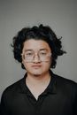 A focused portrait of a young man with curly hair and glasses, wearing a black shirt.